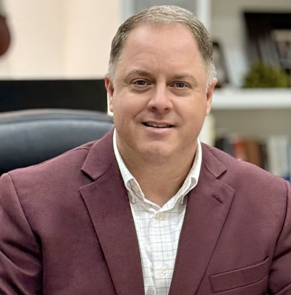 Man in a burgundy blazer and white collared shirt smiles in an office setting.