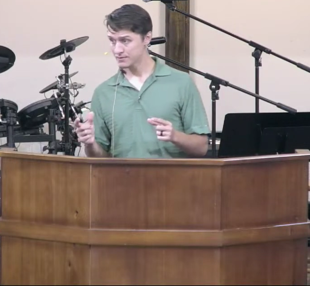 Man in green shirt speaks at a wooden podium, mic in front. Drum set in background.