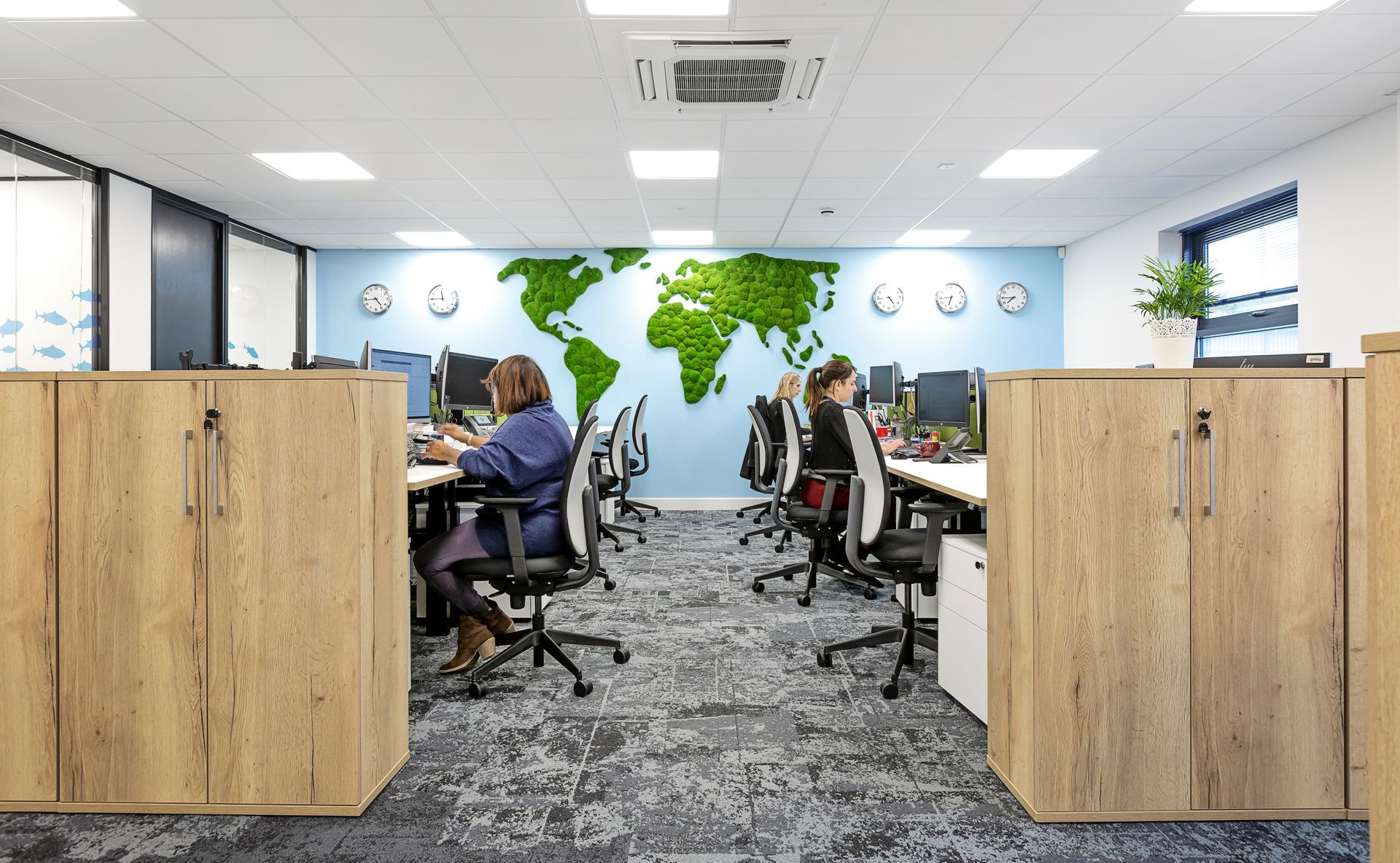 A group of people are sitting at desks in an office with a map of the world on the wall.