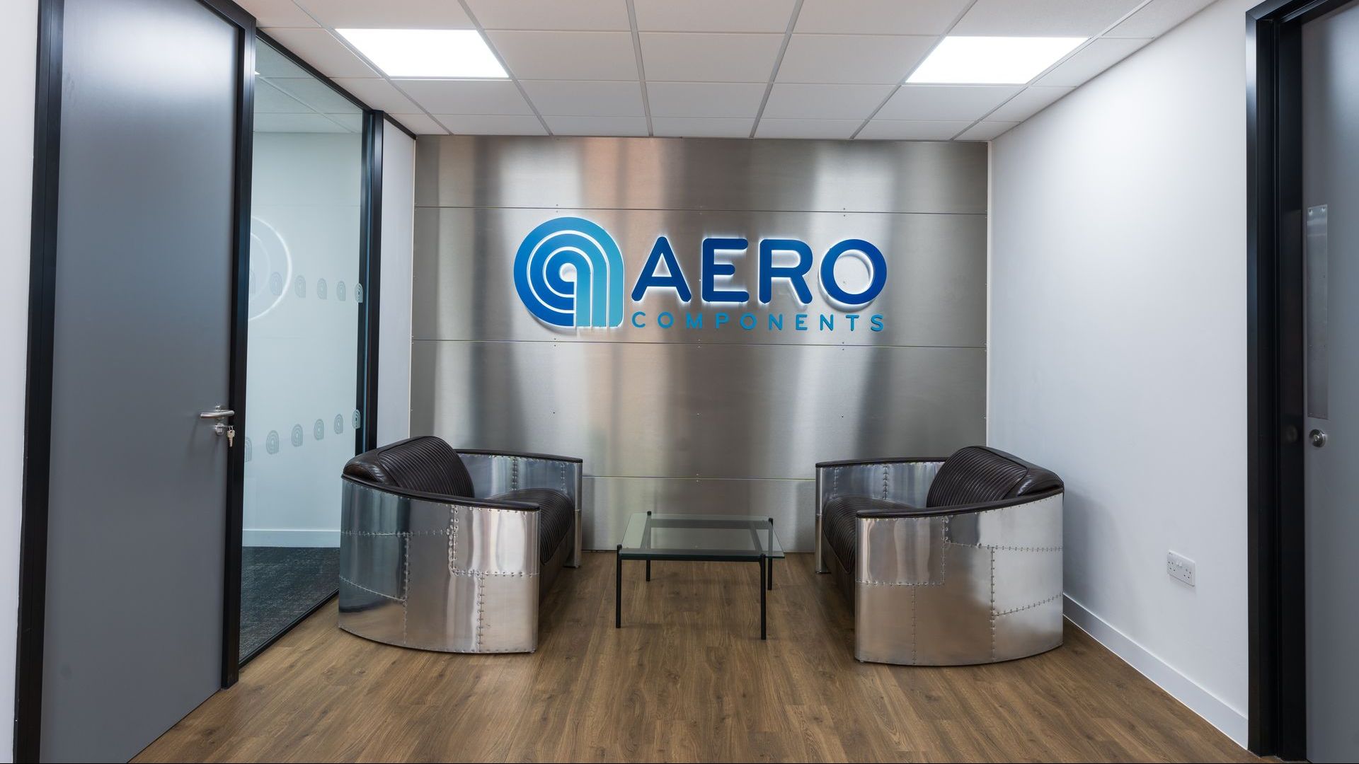 Waiting area with metal chairs, glass table, logo on a silver wall, and a wood floor.