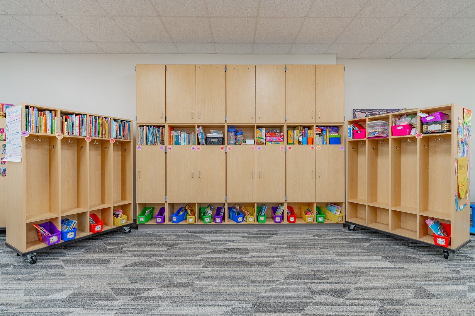 Light-wood classroom storage units and lockers with colorful bins against a gray carpeted wall.