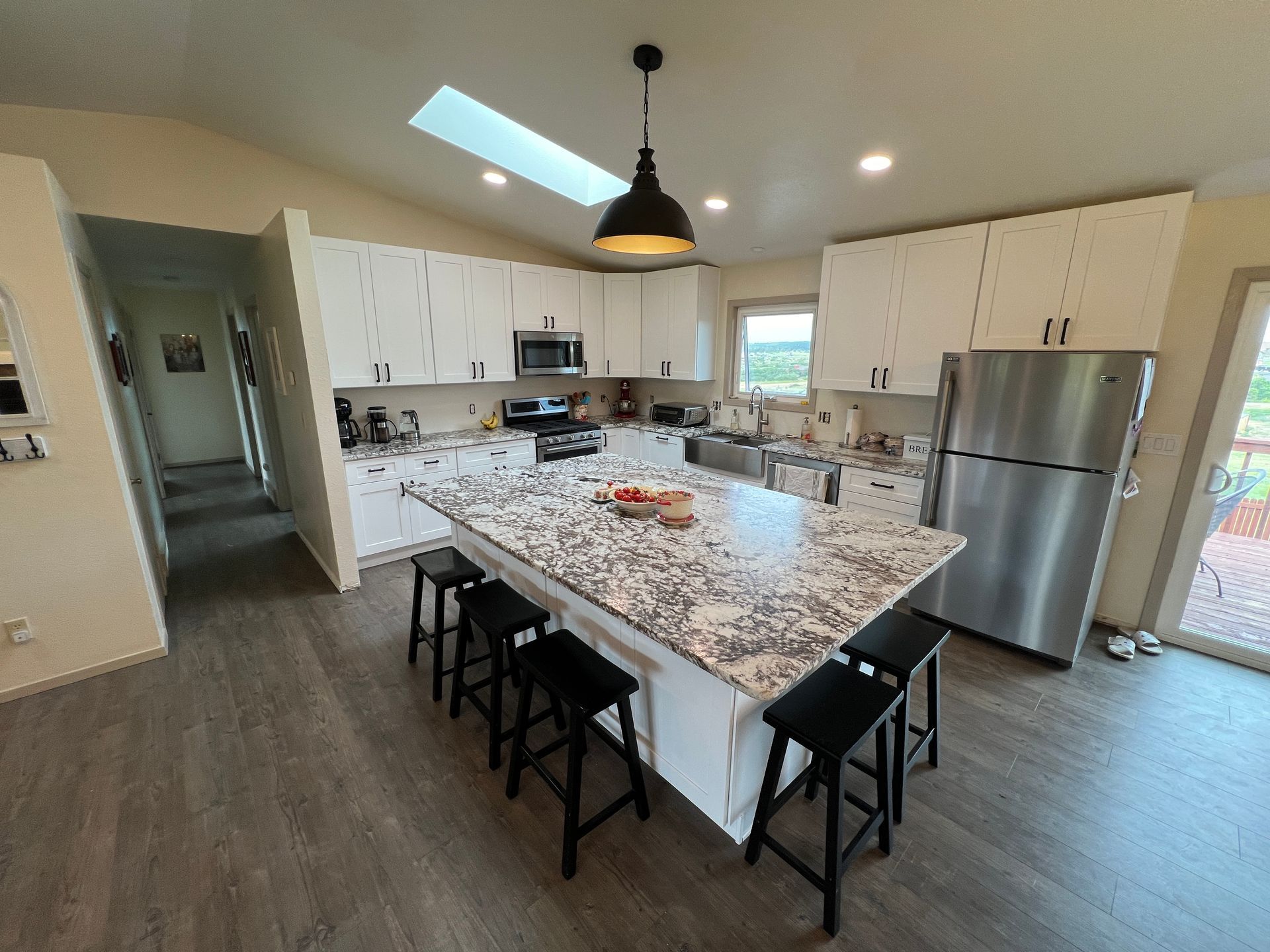 Modern white kitchen with a granite-topped island, four black stools, stainless steel appliances, and a skylight.