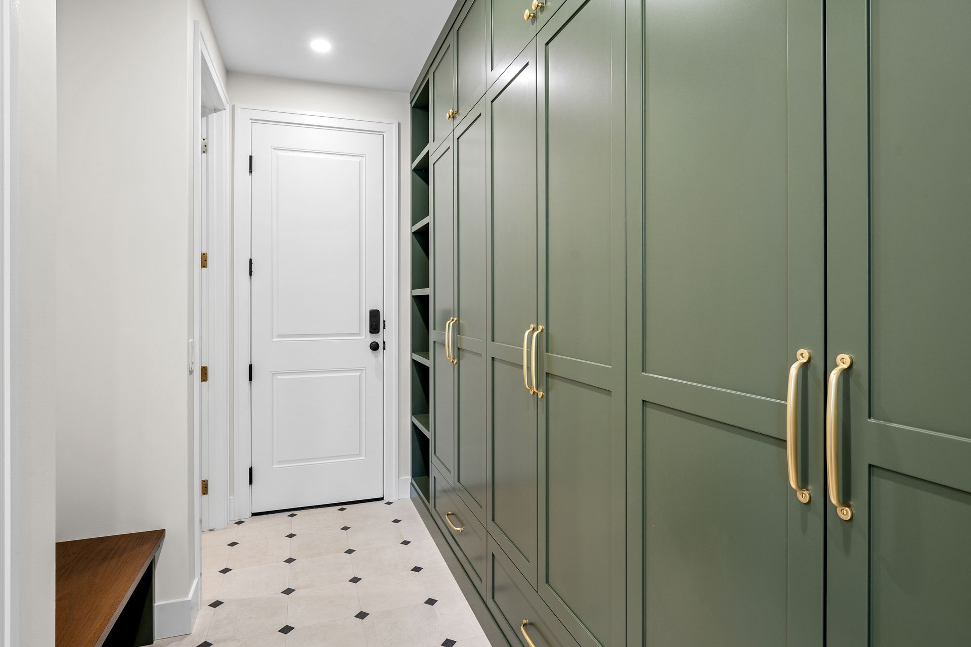 A hallway featuring sage green floor-to-ceiling cabinets with gold handles, white walls, and a white door with black accents.