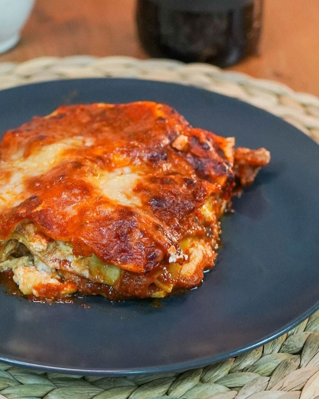 Una fetta di lasagna al forno con formaggio, adagiata su un piatto scuro e ricoperta da una salsa di pomodoro dorata.