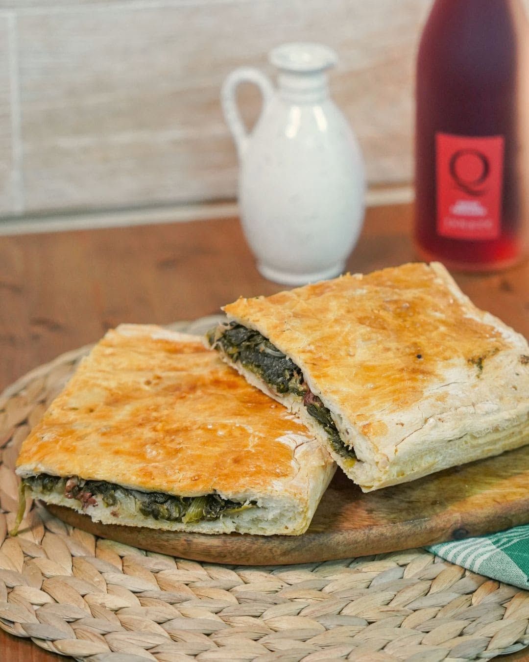 Fette di pasta sfoglia salata dorata su un tagliere di legno, con una brocca bianca e una bottiglia rossa sullo sfondo.