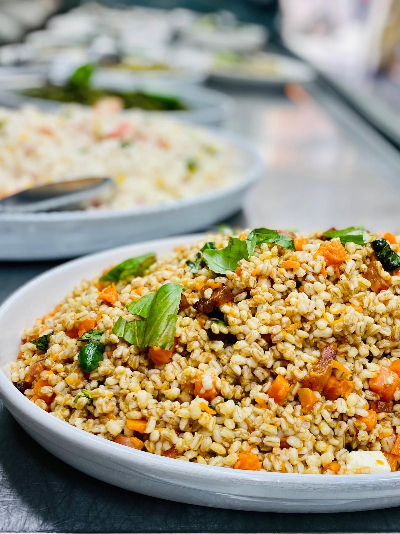 Una ciotola di couscous guarnita con verdure ed erbe aromatiche su un tavolo, con un piatto sfocato sullo sfondo.