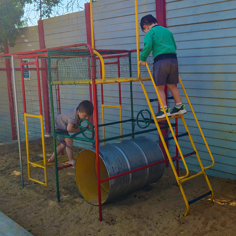 Two children playing on a colorful playground structure with a barrel in a sand pit.