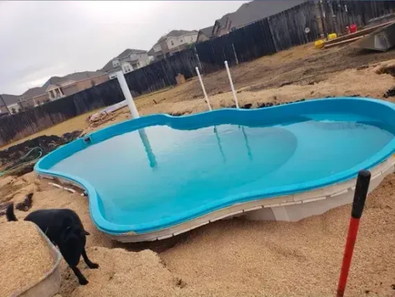 A bright blue, kidney-shaped swimming pool shell sits in a sandy backyard under construction, with a dog nearby.