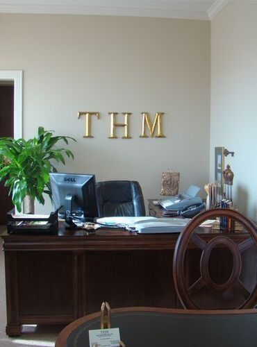 An office desk with a computer, leather chair, and the letters THM on the wall.