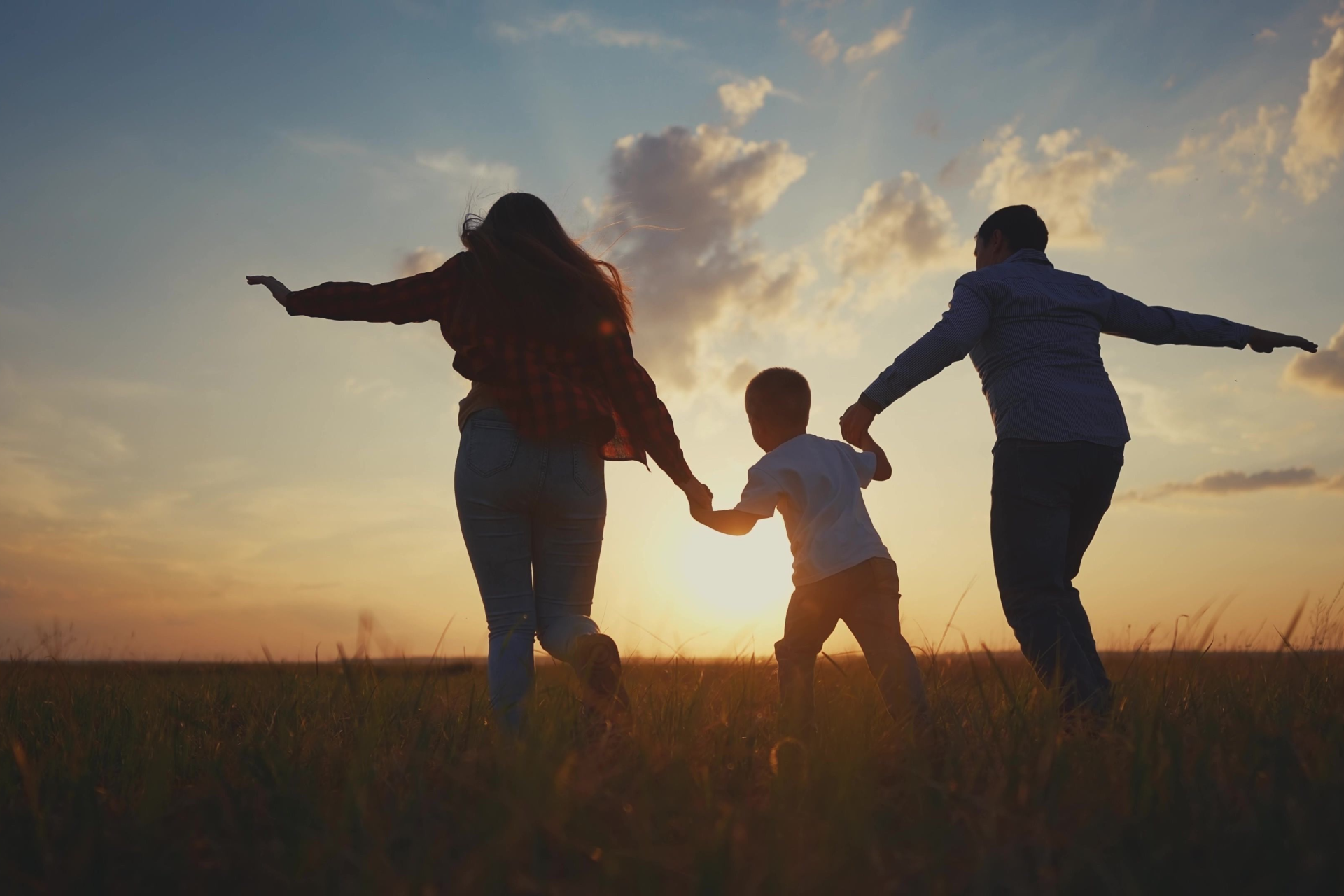 Family holding hands, silhouetted running in field at sunset.