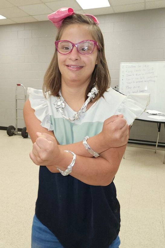 A person wearing a pink bow, glasses, and aluminum foil jewelry, holding their arms crossed in front of them.
