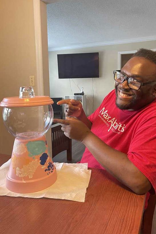 A person smiling while pointing to a DIY gumball machine made from a painted terracotta pot and a glass bowl.