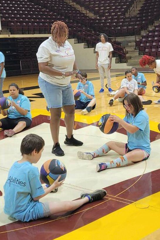 An instructor guides people sitting on a basketball court floor holding multicolored basketballs.