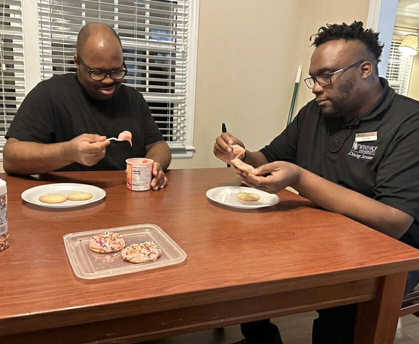 Two people sit at a wooden table icing plain cookies with pink frosting from a tub, with decorated cookies nearby. Residential Support York County SC