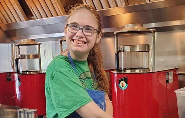A smiling person in a green shirt and blue apron standing in a kitchen between two red coffee brewers.