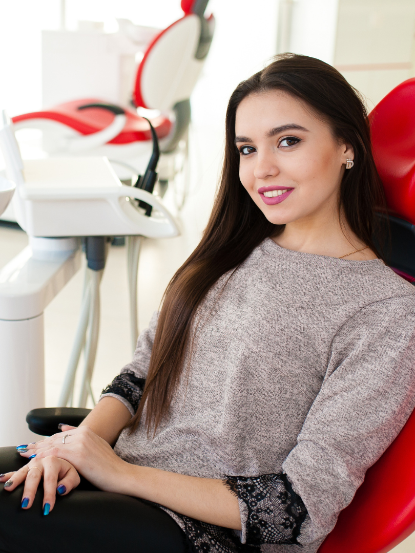A woman is sitting in a red dental chair in a dental office.
