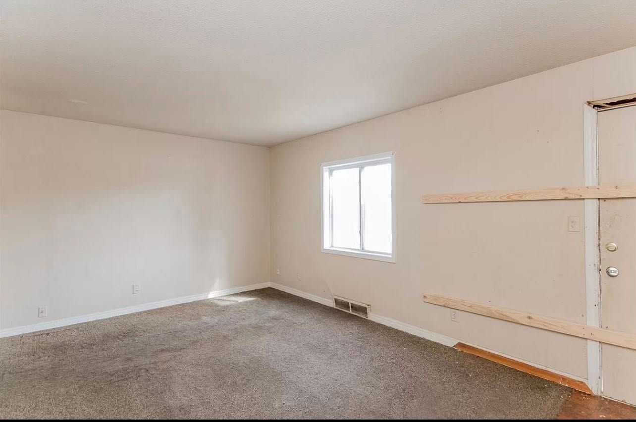 Empty room with brown carpet, window, and boarded-up door. Beige walls and white ceiling.