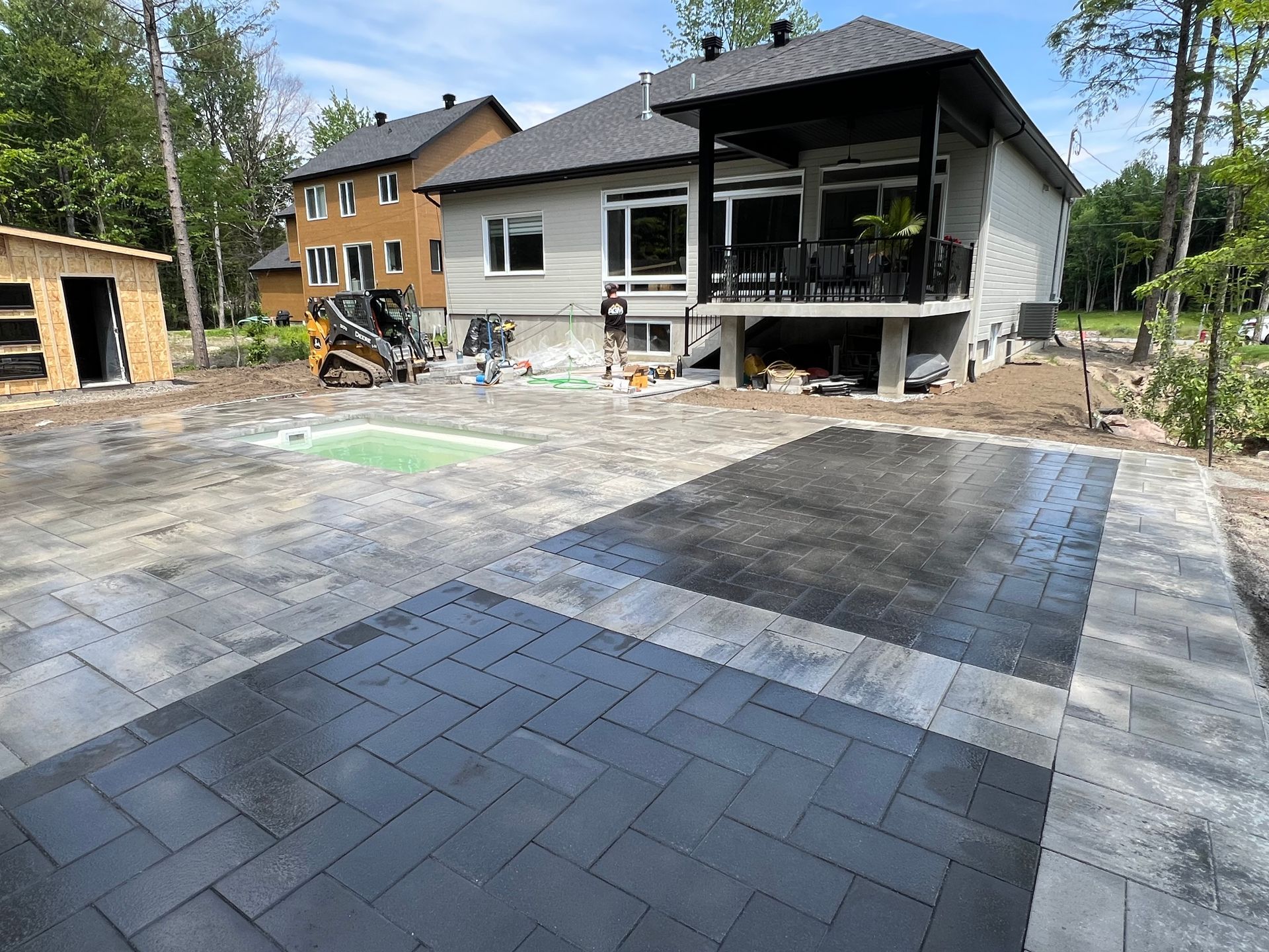 Une nouvelle terrasse en pierre grise et noire est en construction dans le jardin d'une maison moderne, avec une petite piscine à proximité.