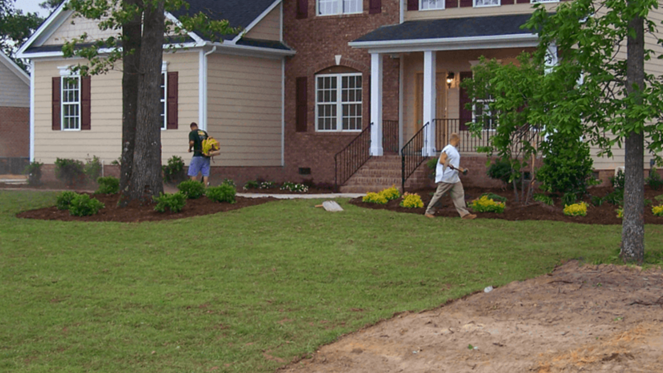 Two people spraying a lawn in front of a brick and tan house in Wilmington, NC.