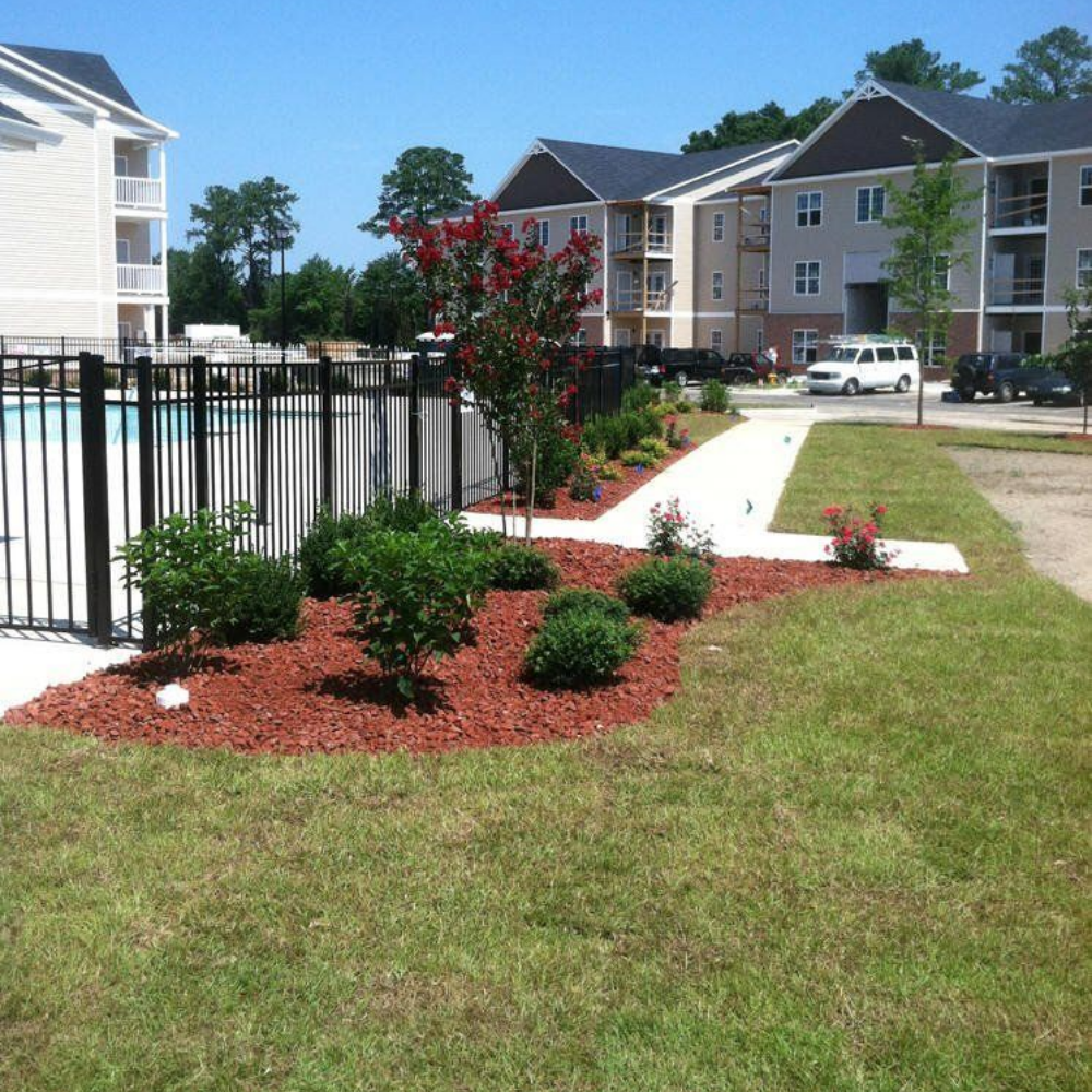 A landscaped area with red mulch and small shrubs by a metal pool fence, beside a sidewalk and apartment buildings in Raeford, NC.