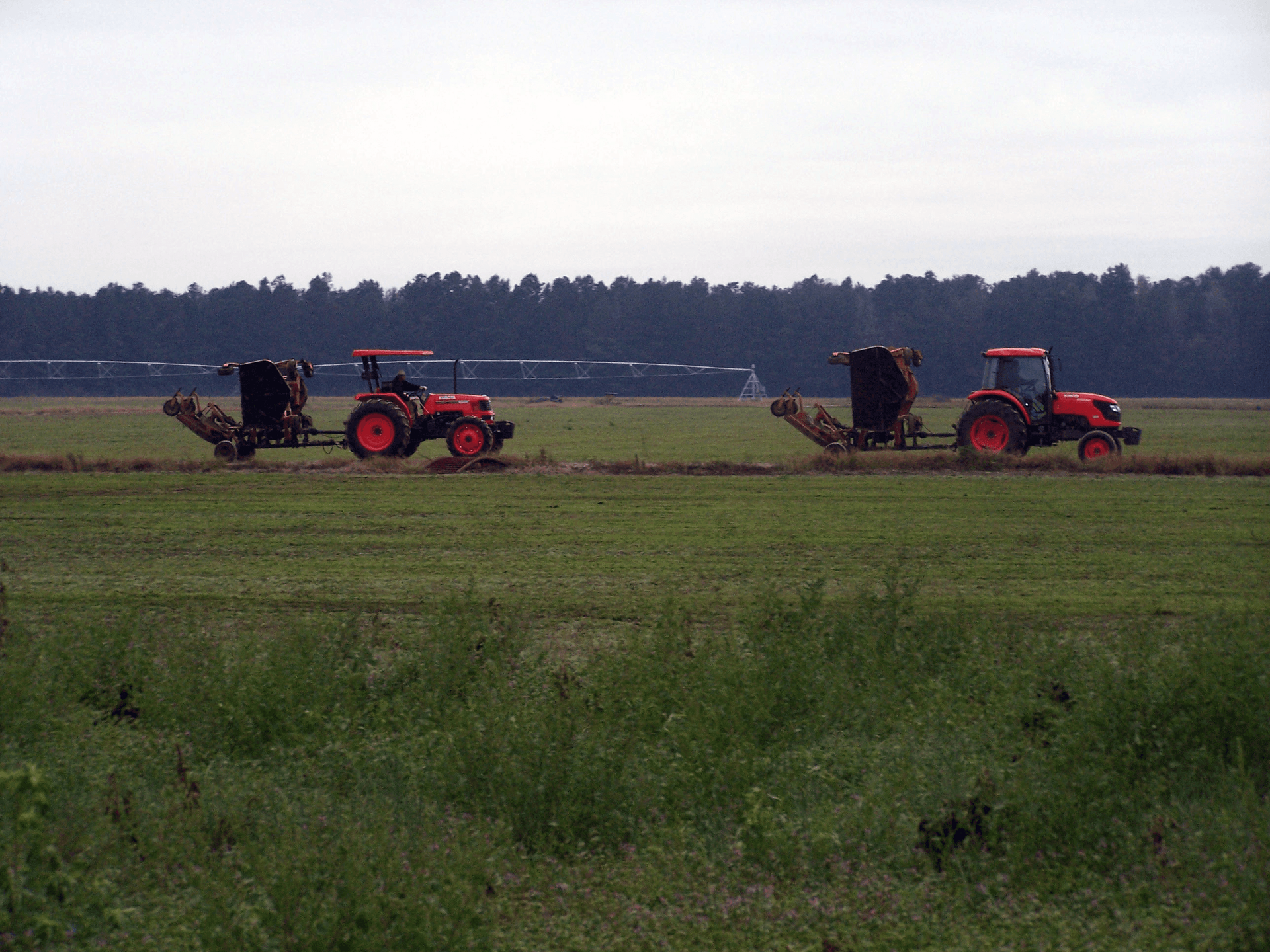 Two tractors are working in a field with trees in the background.