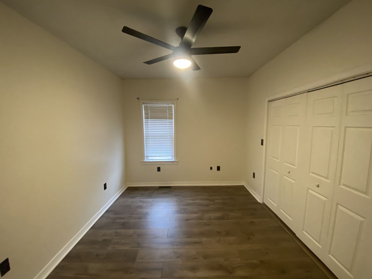 Empty bedroom with wood floors, white walls, closed closet doors, and ceiling fan.