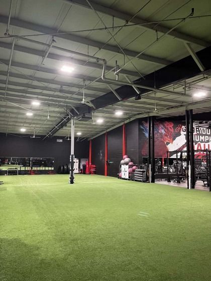 Gym interior with green turf, black walls, and equipment. A person stands near weights.