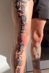 Tattooed lower legs of a person standing indoors, showing black script on one calf and a portrait tattoo on the other