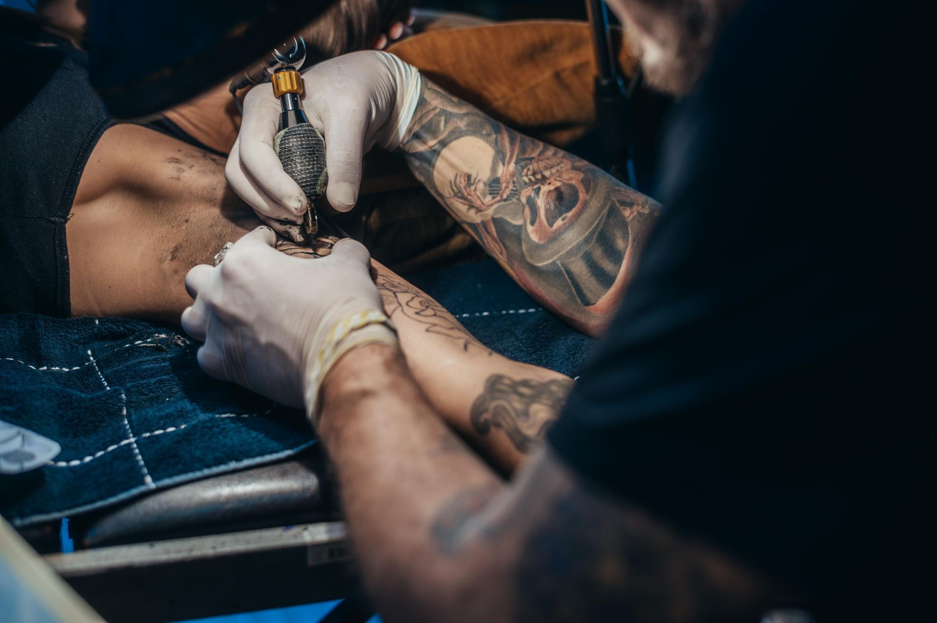 Person Getting A Tattoo On Their Arm From An Artist Wearing Gloves; Blue Table — Tattoo Inc Studio in Burleigh, QLD