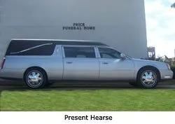 Silver hearse parked on grass beside a wall, with “Present Hearse” caption below and “Price Funeral Home” sign above.