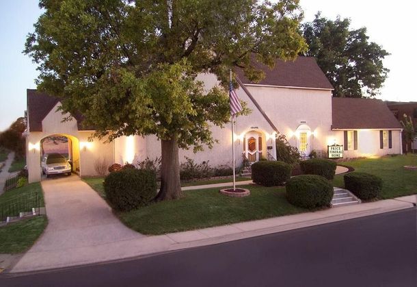Single-story suburban home at dusk with warm porch lights, large tree, driveway, and trimmed front lawn