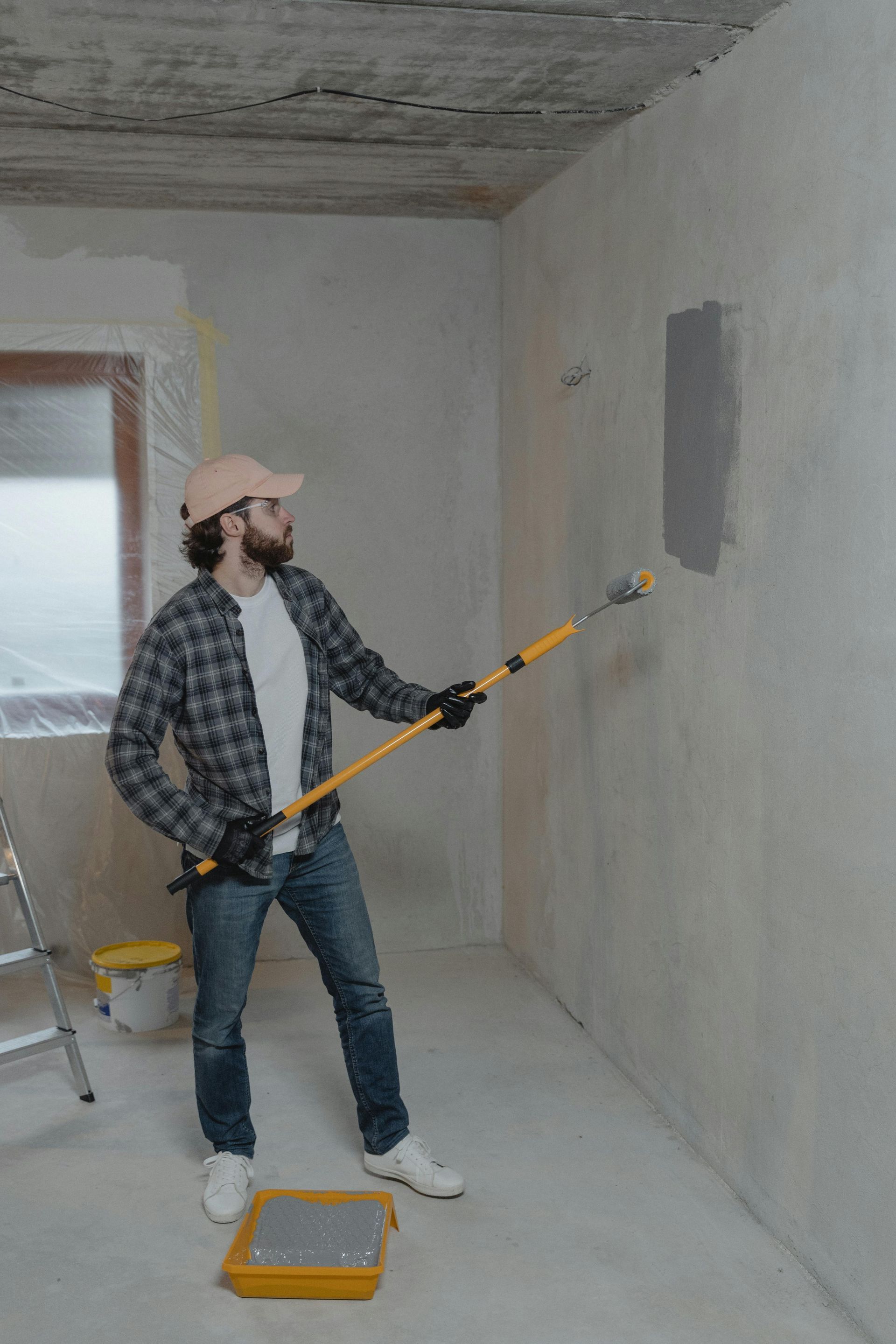 A person wearing a plaid shirt and cap uses a long-handled roller to paint a gray patch on an unfinished white wall.