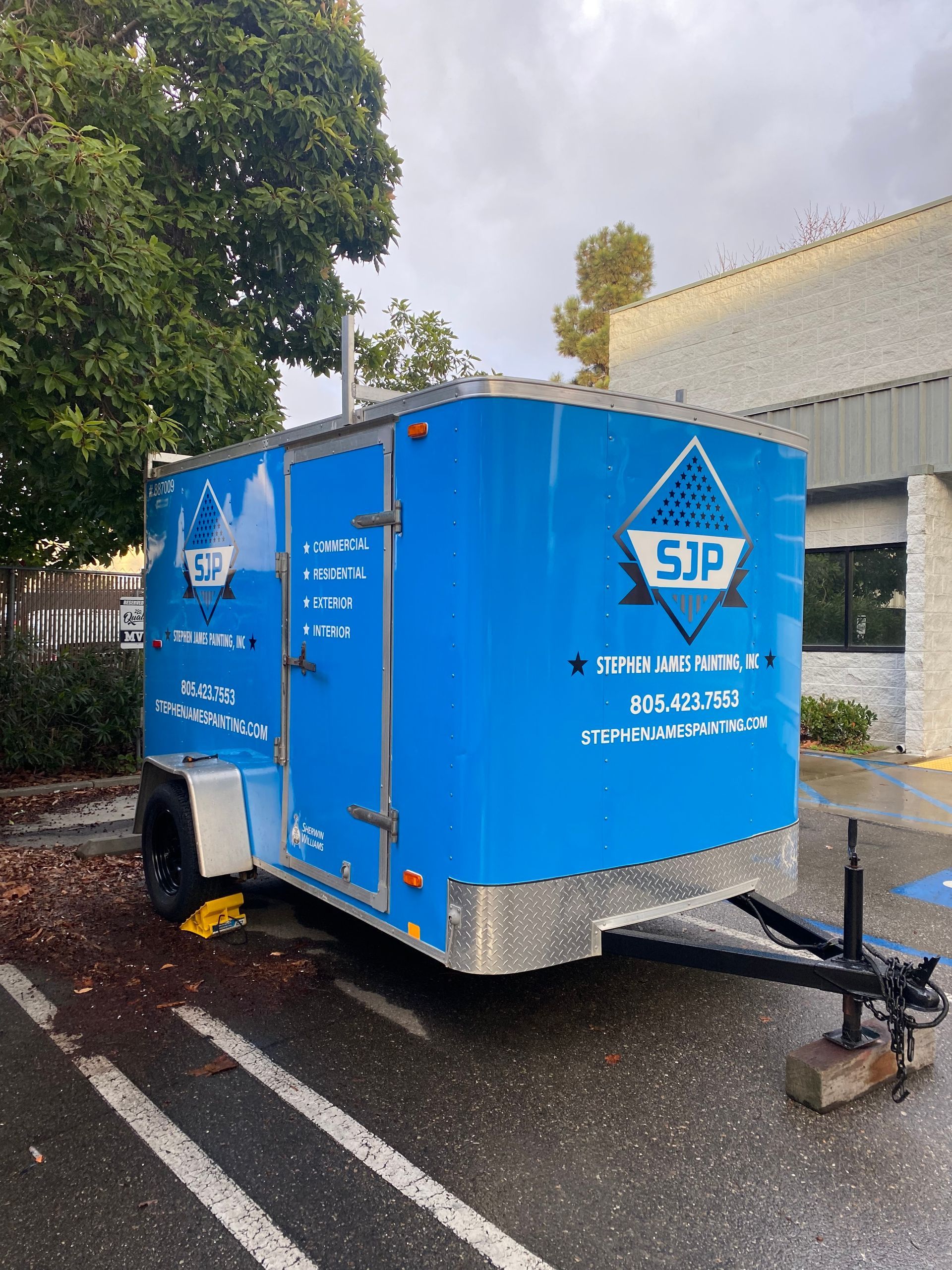 Blue enclosed utility trailer parked outdoors on wet pavement beside a building and trees