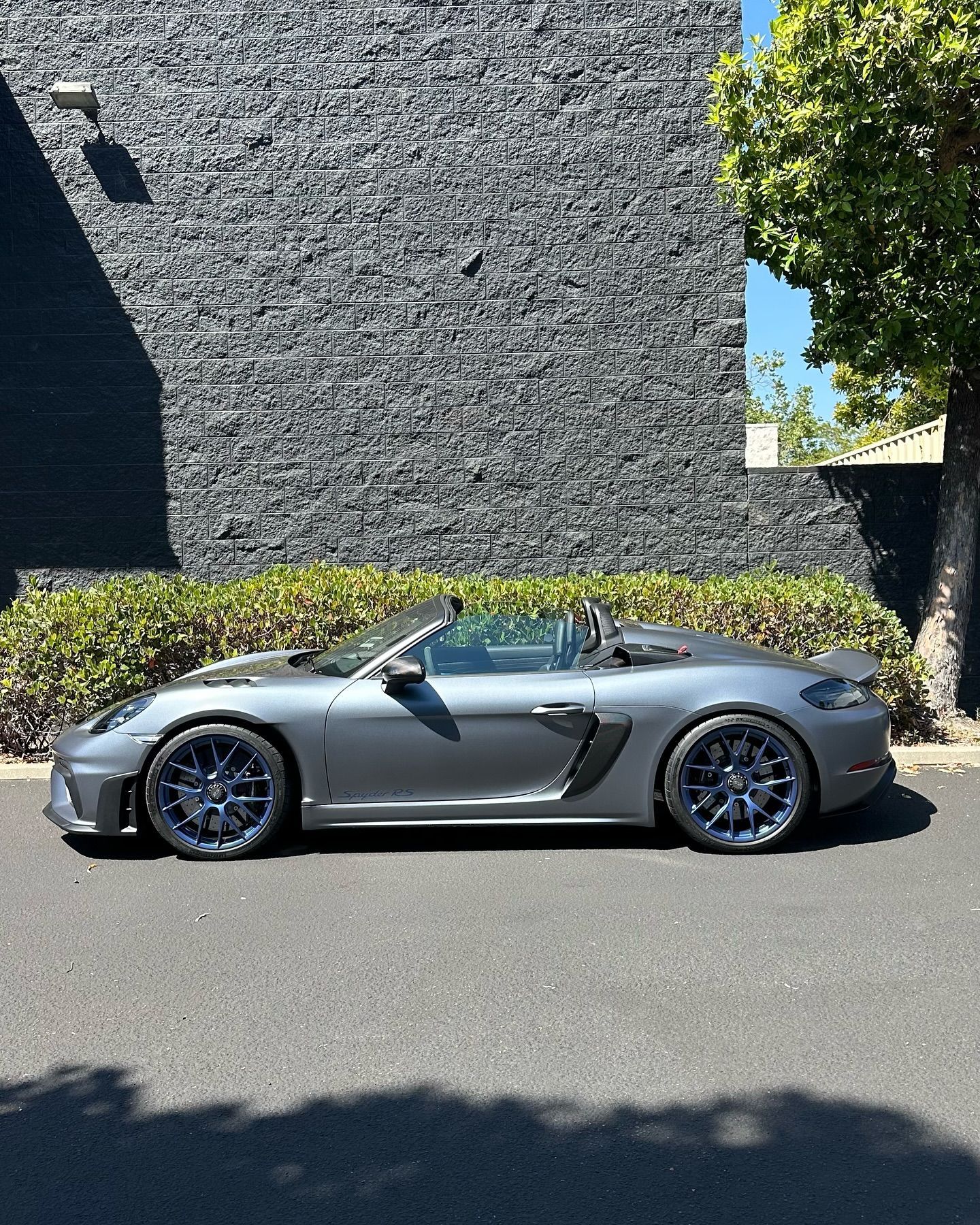 Gray convertible sports car parked beside a dark stone wall in sunlight