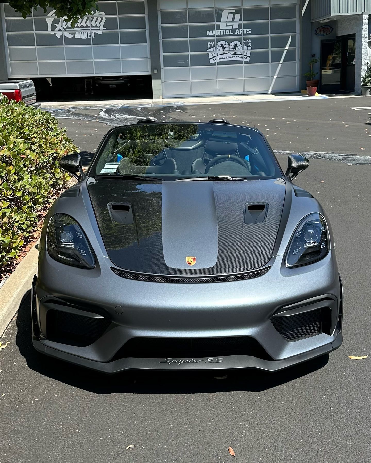 Gray sports car parked outside a storefront, front view with a black hood and open roof.