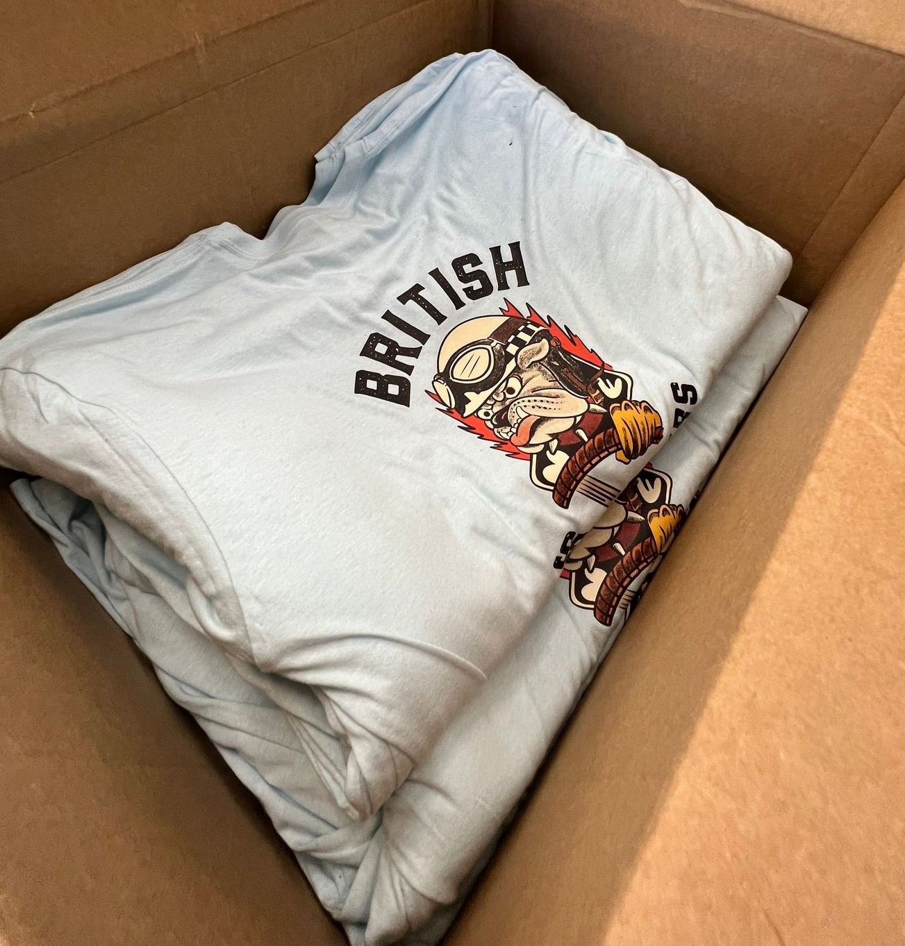 Folded white T-shirt with “BRITISH” graphic, packed inside a cardboard box.