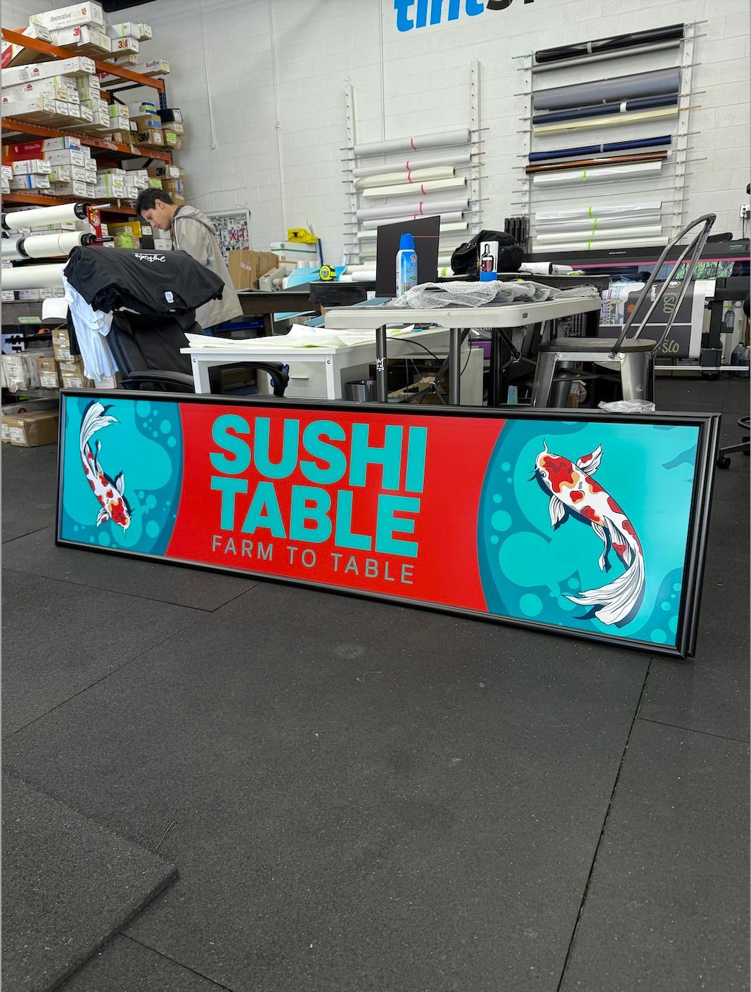 Bright turquoise sushi table display with a person working behind it in a warehouse-like space.