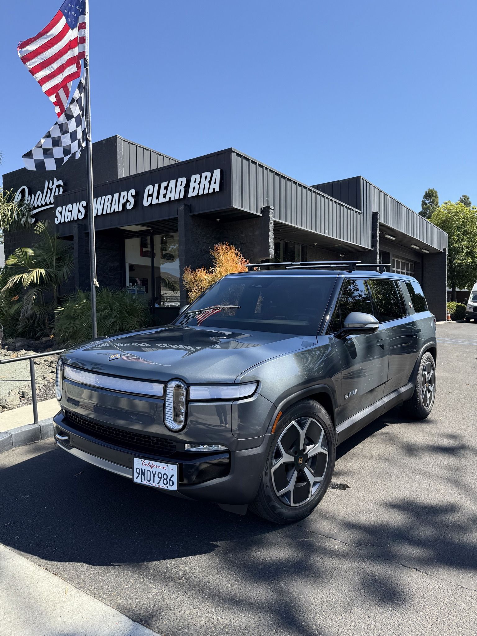 Gray SUV parked outside a building with a waving American flag under a clear blue sky.