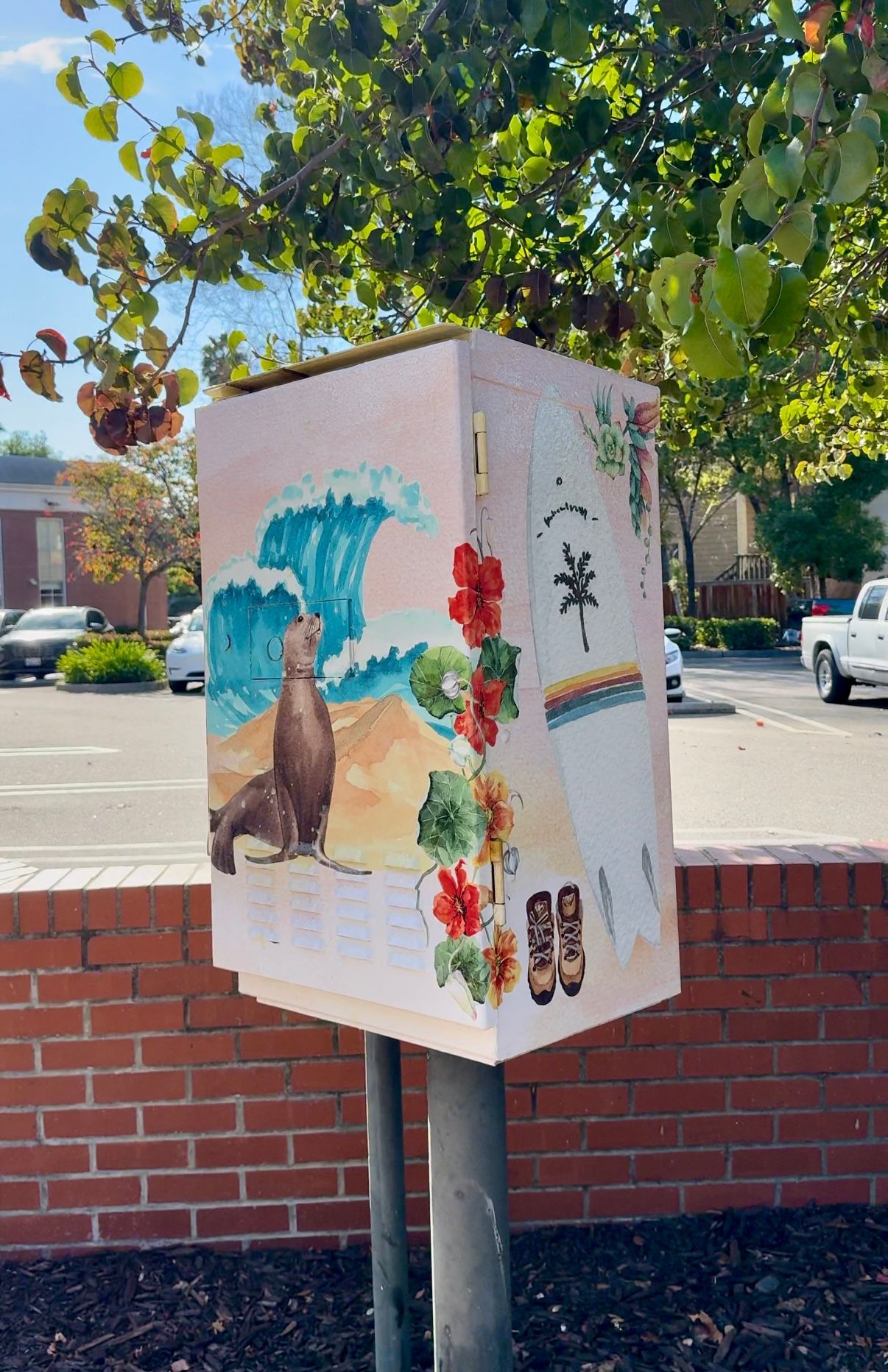 Decorated utility box with colorful murals beside a brick wall under trees in a parking lot