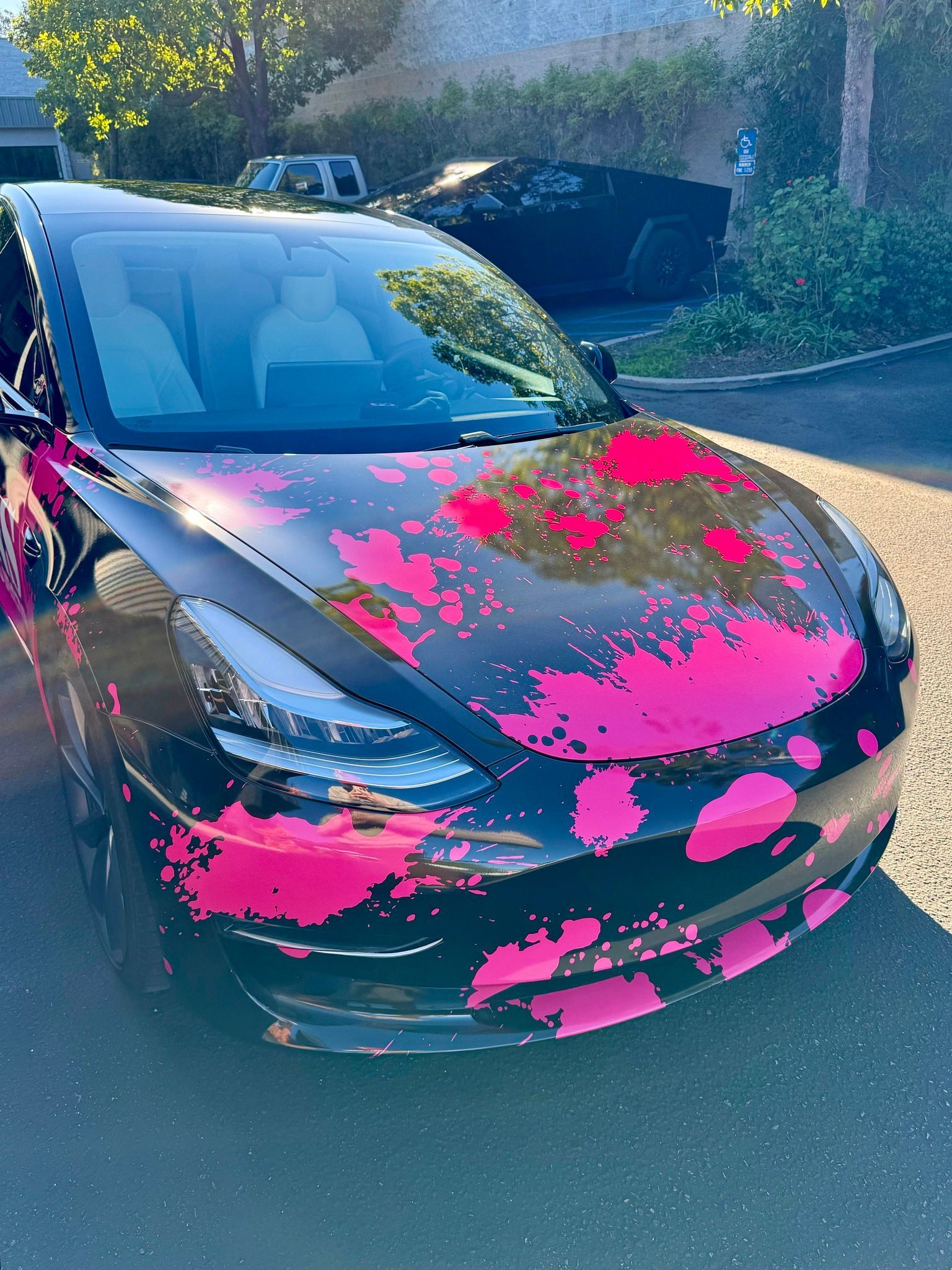 Black car with bright pink paint splatter on the hood, parked outdoors in sunlight