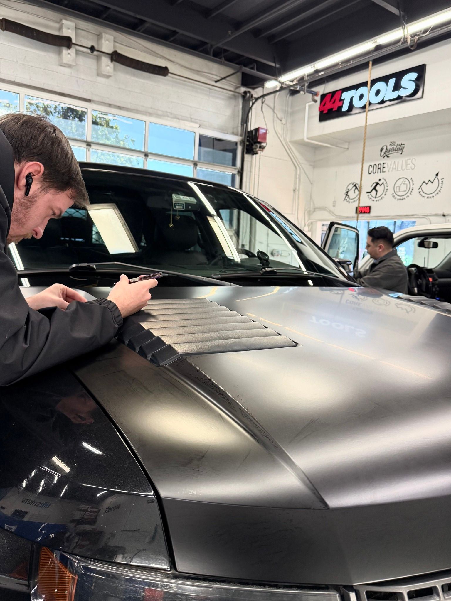 Matte black car being detailed in an auto shop, with a technician polishing the hood.