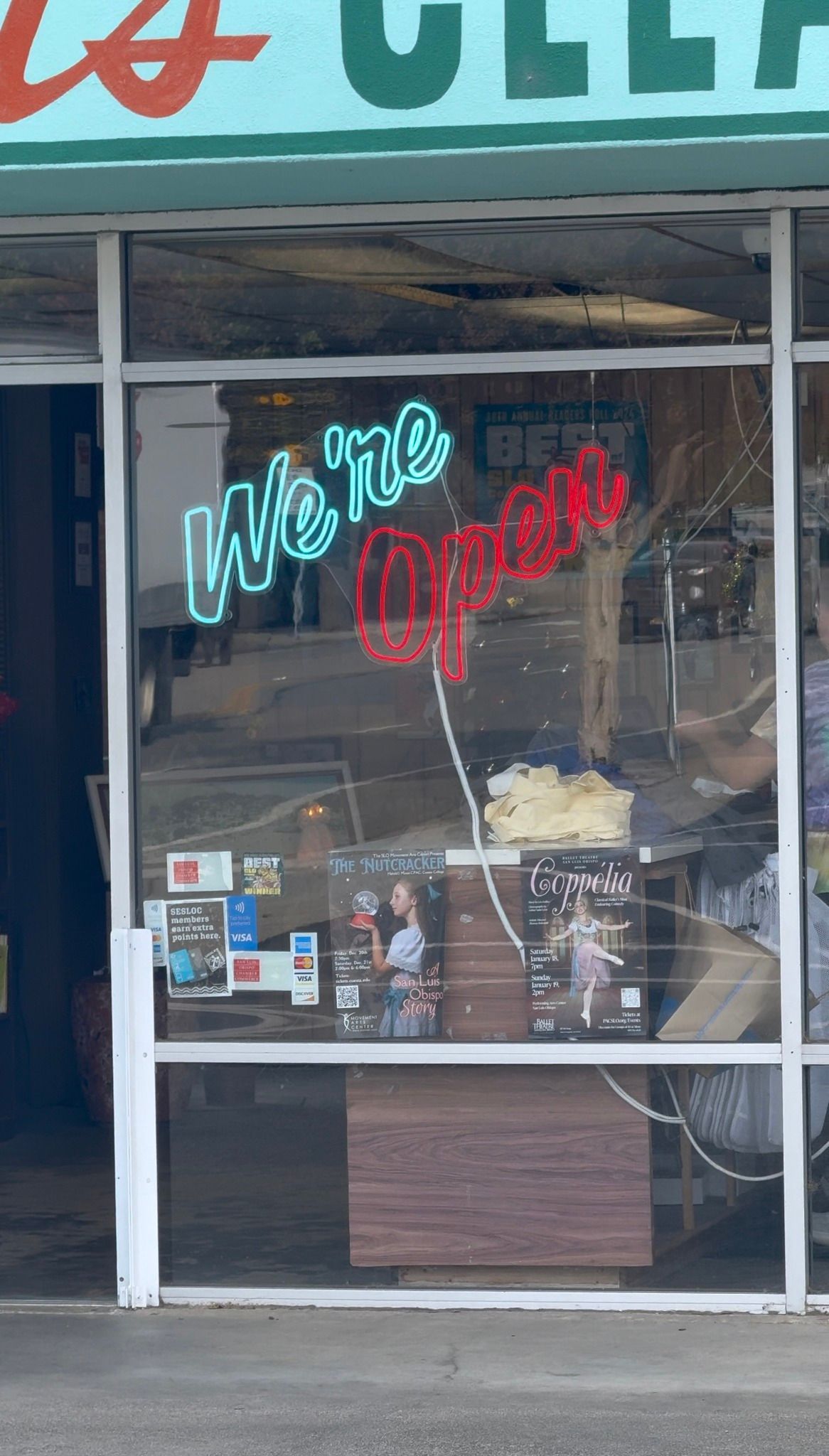 Storefront with a glass door and neon “We open” sign in turquoise and red, with items visible inside