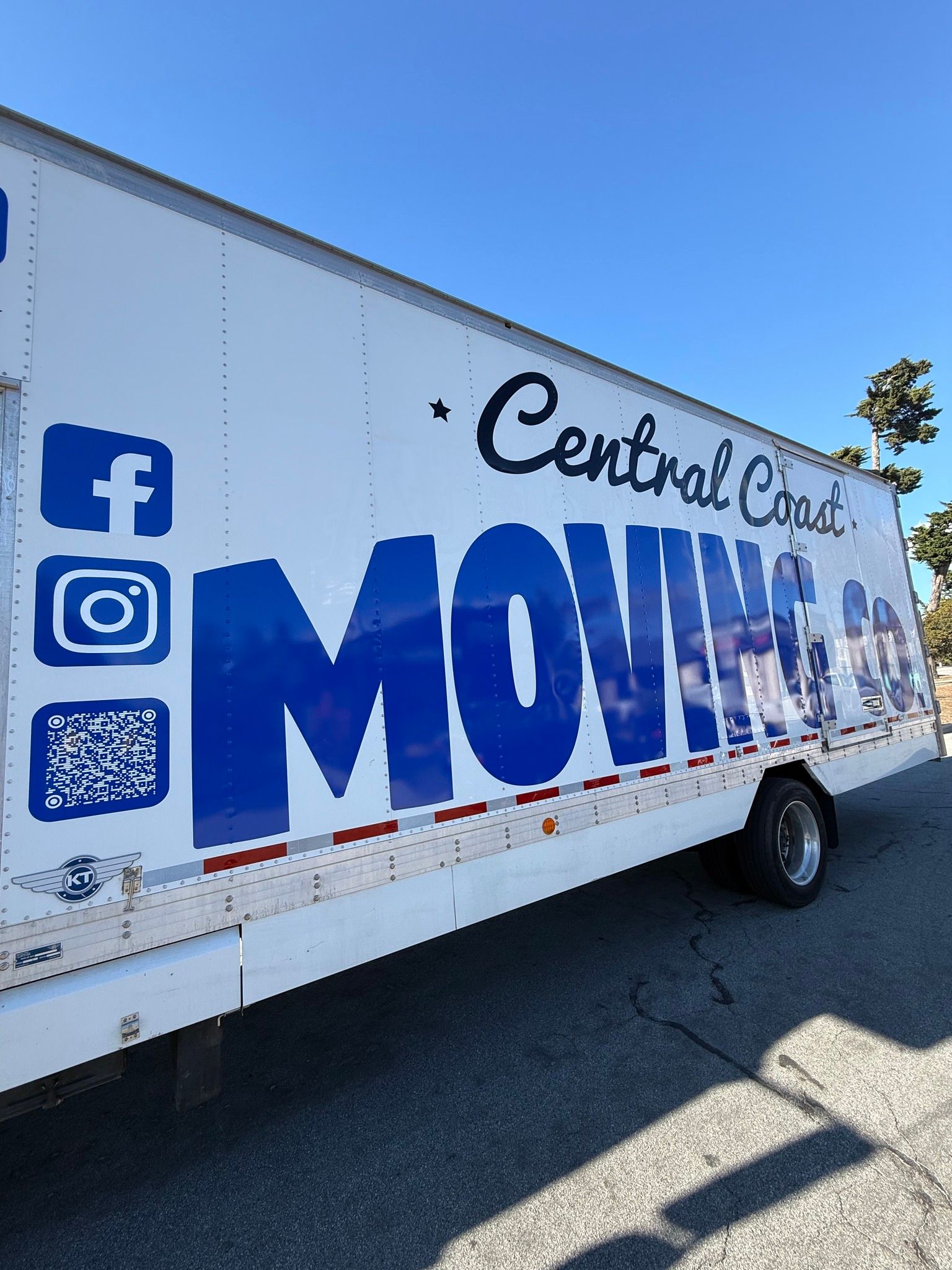White moving truck with blue “MOVING” lettering and social media icons on the side in a sunny parking lot
