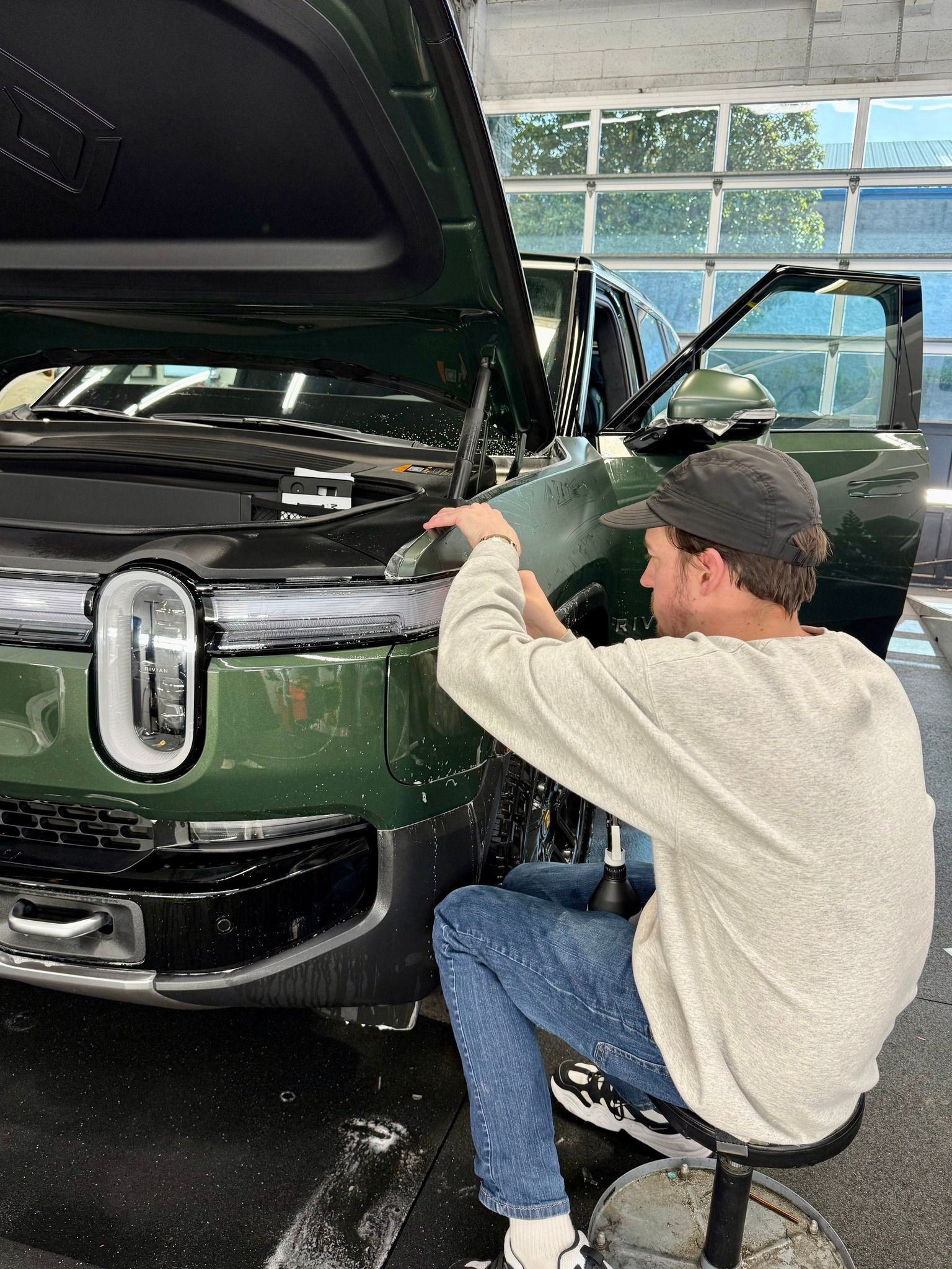 Man cleaning a dark green SUV at a car wash, with hood open and water spraying nearby.