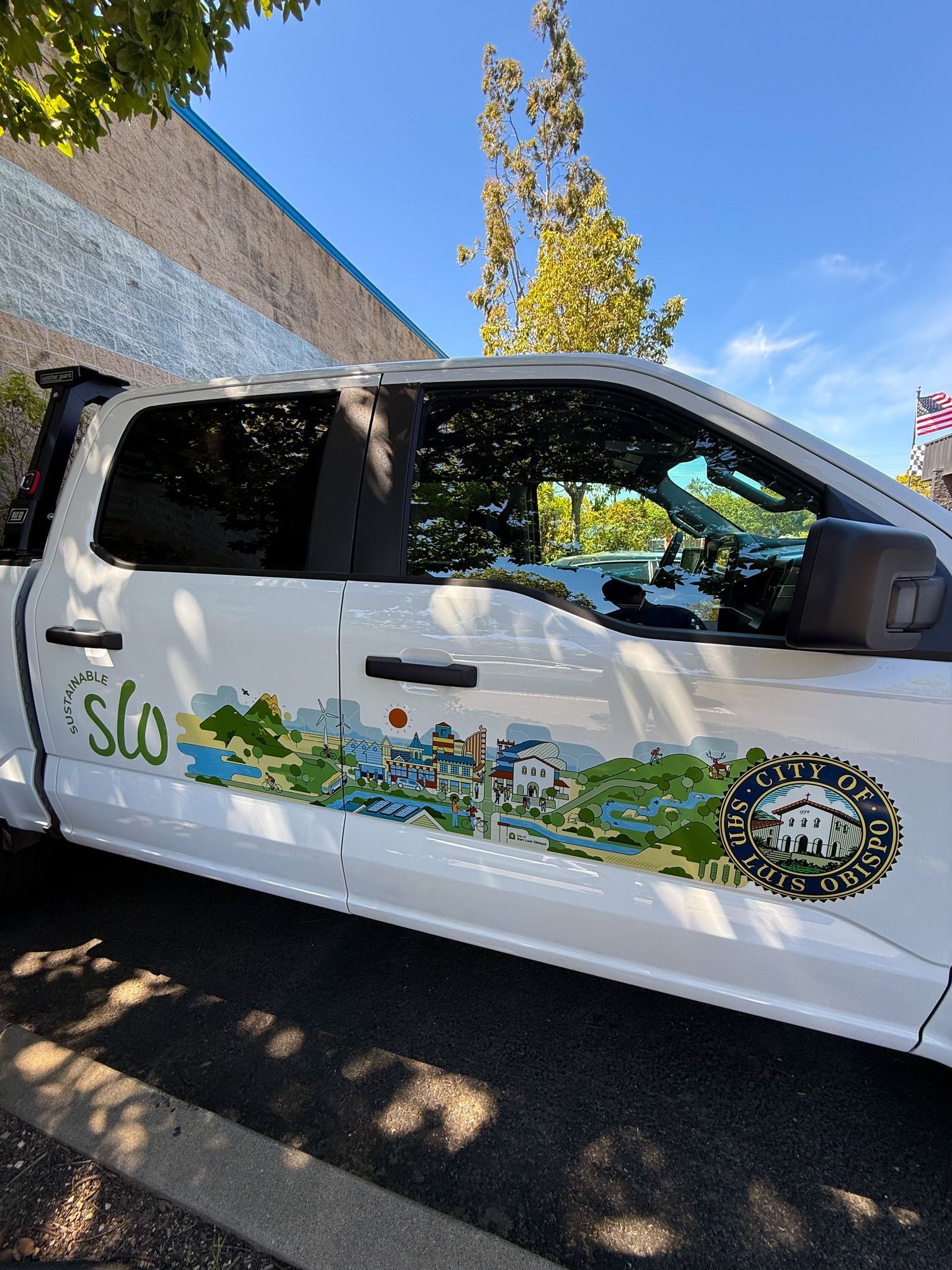 White pickup truck with colorful SLO nature mural parked on a sunny street near a stone wall.