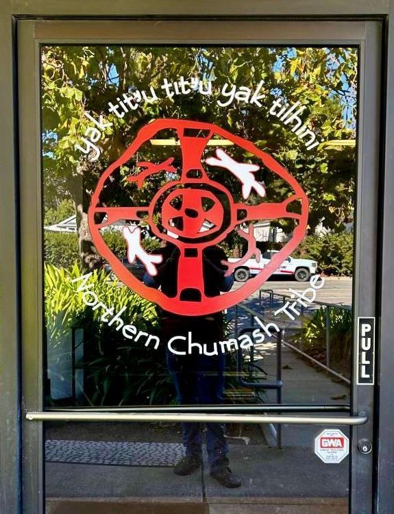 Glass door with red circular emblem and white lettering, reflecting trees outside.