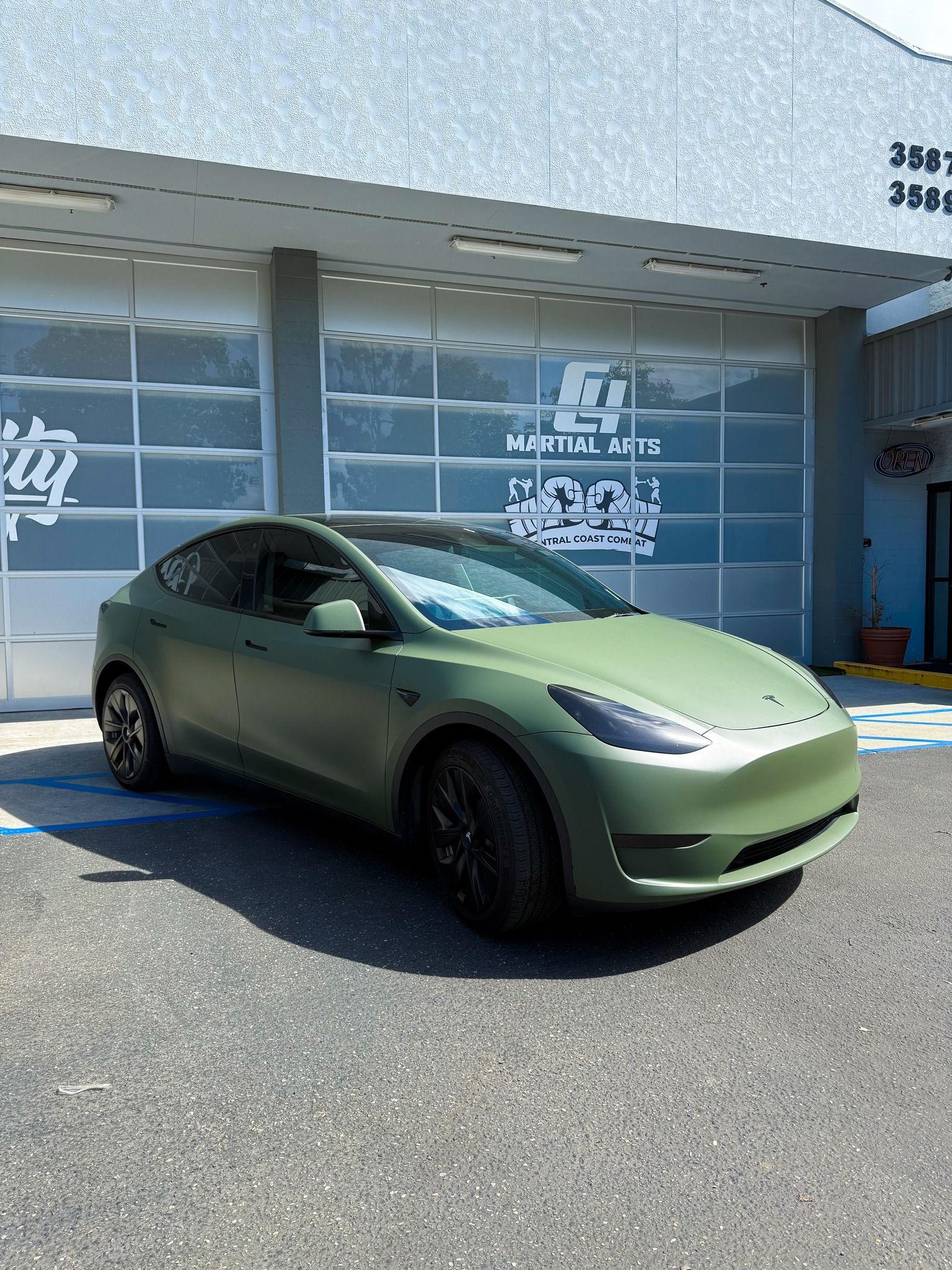Matte green sedan parked outside a storefront with large glass windows in bright sunlight