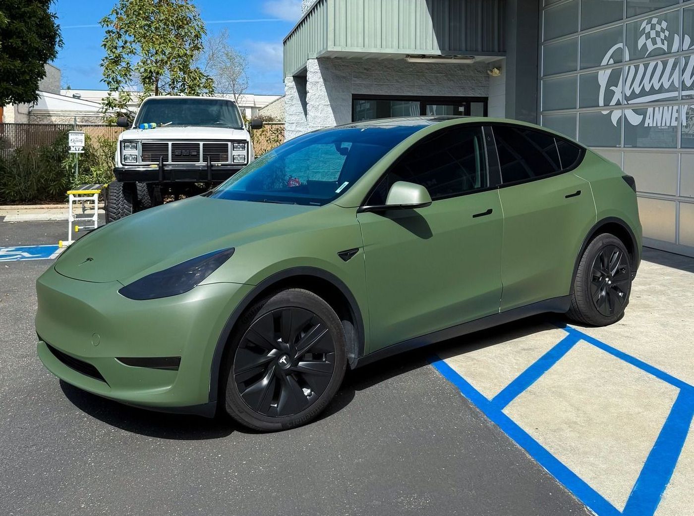 Matte green Tesla parked in a blue-striped accessible parking space outside a building