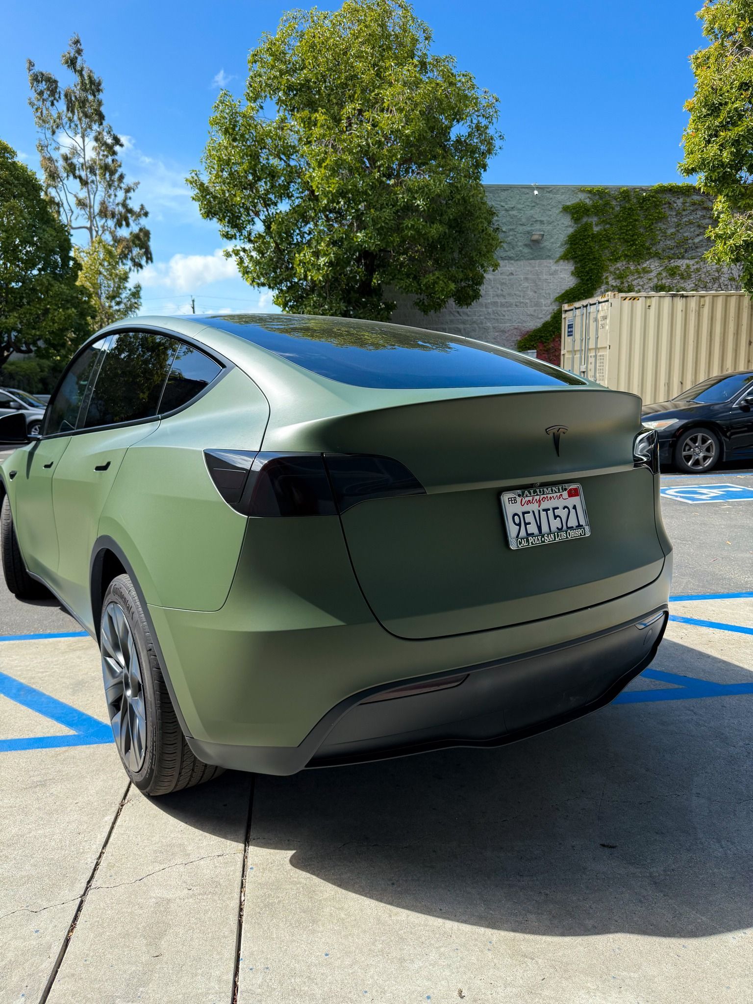 Matte olive-green Tesla parked in a sunny lot with trees and blue sky