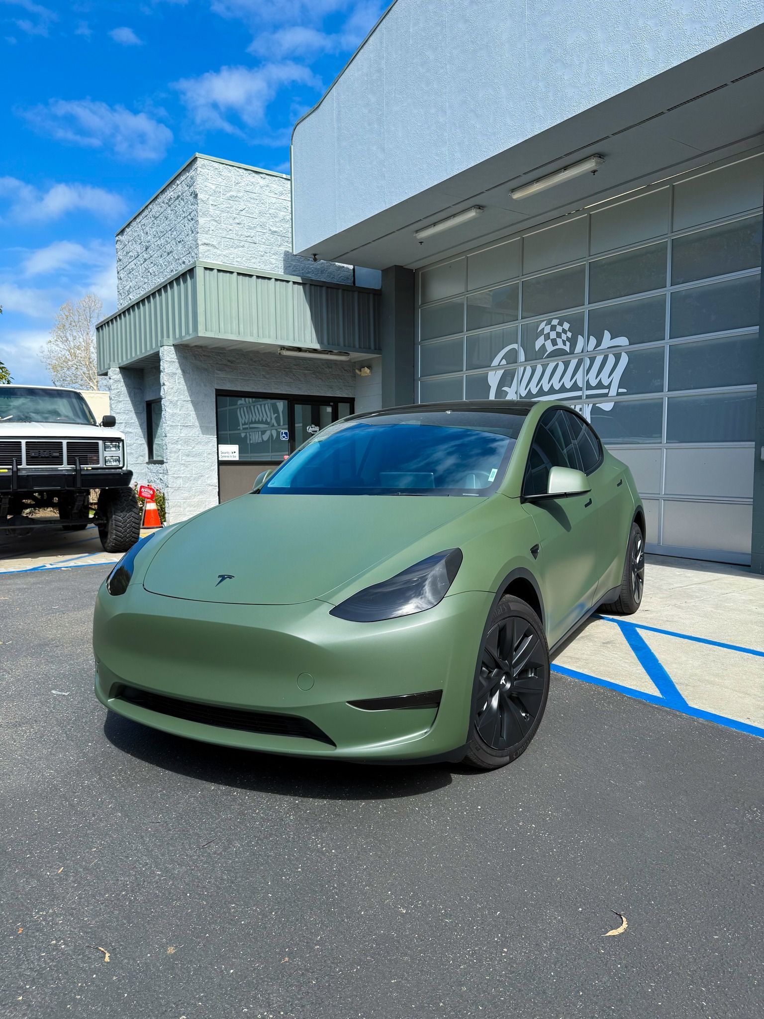 Matte green Tesla parked outside a modern building on a sunny day.
